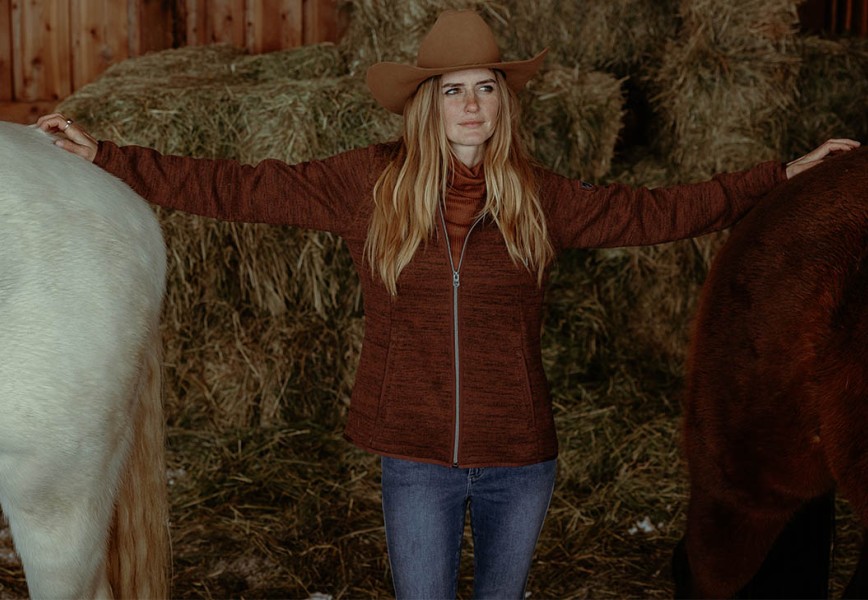 A woman stands between two stabled horses in womens fall jacket