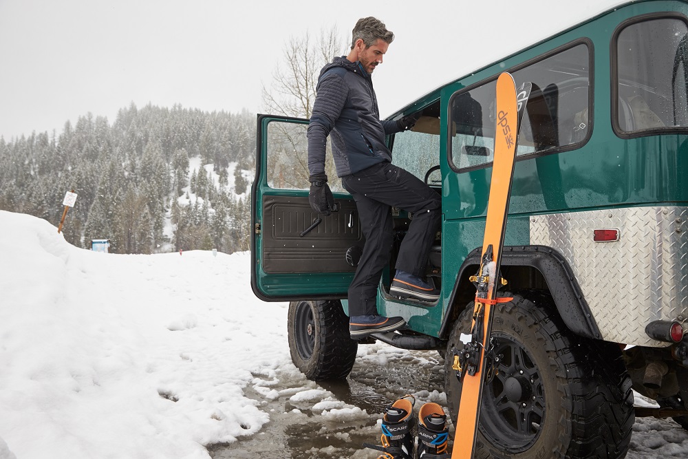 A man standing on a van with a pair of skis placed on the back door, snowy background.