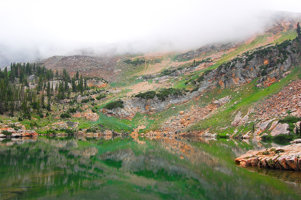 fog on top of mountain with green lake in front