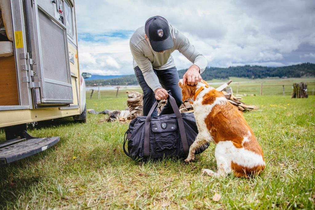 man in KÜHL cap holding KÜHL bag petting white and orange dog