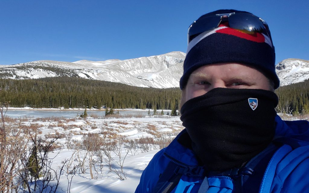 man wearing KÜHL neck gaiter facing camera on snowy field during daytime